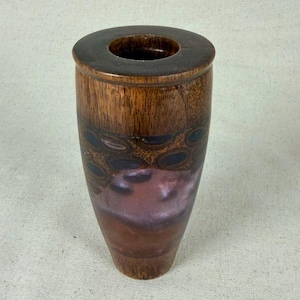 Może przedstawiać: Ręcznie wykonany drewniany wazon o cylindrycznym kształcie. Wazon ma ciemnobrązową obręcz i podstawę, z jaśniejszym wzorem słojów drewna na korpusie. Ozdobne ciemnoniebieskie i czarne owalne kształty są namalowane na górnej części wazonu, z gradientem fioletu i brązu na dolnej połowie.