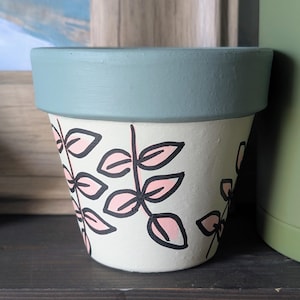 May include: A small, hand-painted ceramic flower pot. The pot is off-white with a light blue rim. Pink leaves with black outlines decorate the pot's exterior. The pot is designed for indoor or outdoor use.