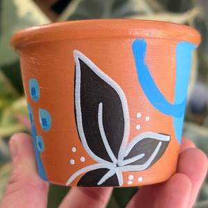 May include: A small, orange terracotta plant pot with abstract painted designs. The pot features a large black leaf with white outlines, blue brushstrokes, and small blue and white dots. The pot is held in a hand.