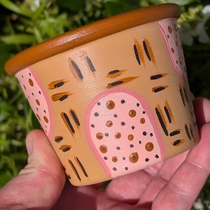 May include: Hand-painted ceramic pot with a tan base, brown rim, and pink and brown abstract designs. The pot features pink arched shapes with brown dots and brown vertical and horizontal brushstrokes. A decorative item for plants.