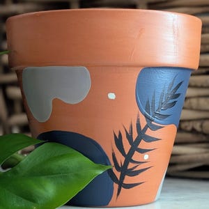 May include: Terracotta-colored ceramic plant pot with abstract painted design. The design includes gray, navy blue, and white shapes, with a black fern-like illustration. A green leaf is in the foreground.