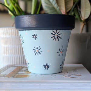 May include: A hand-painted, light blue flower pot with a dark blue rim. The pot is decorated with small, stylized flowers in dark blue and peach. The pot is sitting on a stack of books, with green plants in the background.