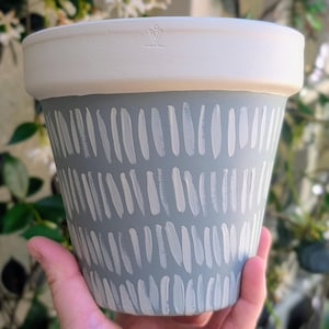May include: A hand-painted ceramic flower pot. The pot features a light gray base with a repeating pattern of white vertical brushstrokes. The rim is painted white. The pot is designed for indoor or outdoor use.