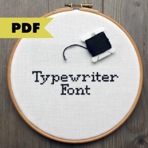 Op de afbeelding: Een witte borduurring met de woorden "Typewriter Font" geborduurd in zwart garen. Een spoel met zwart borduurgaren is inbegrepen.
