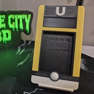 May include: A yellow and black 3D-printed phone case with a white base and a circular button. The case has the letter "U" on top and the text "Designed By MAS FUEGO 3D PRODUCTIONS" on the front. A glowing green "SPACE CITY 3D" sign is in the background.