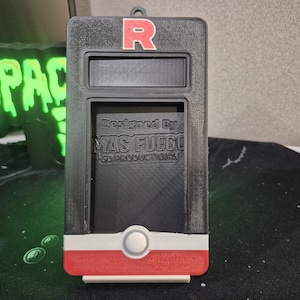 May include: Black and red rectangular case with a white and red accent at the bottom. The top has a red "R" and a rectangular opening. The text "Designed By MAS FUEGO 3D PRODUCTIONS" is visible. A green neon sign that says "SPACE" is in the background.