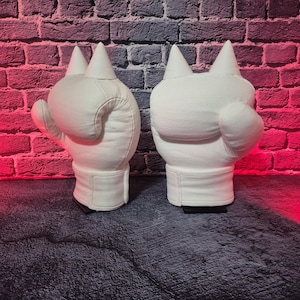 May include: Two white, cat-shaped decorative lamps. Each lamp has a rounded body, a pointed ear, and a rectangular base. The lamps are made of a smooth, matte material and are set against a brick wall with a red and blue gradient.
