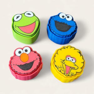May include: Four colorful, character-shaped containers. A green frog, a blue Cookie Monster, a pink Elmo, and a yellow Big Bird. Each container has a cartoon face on the top, with a matching colored base.