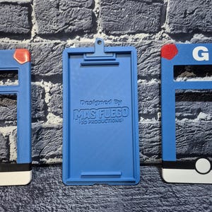 May include: A blue 3D-printed clipboard with a clip at the top and the text "Designed By MAS FUEGO 3D PRODUCTIONS" printed on the front. The clipboard is flanked by two blue frames with red and white accents.