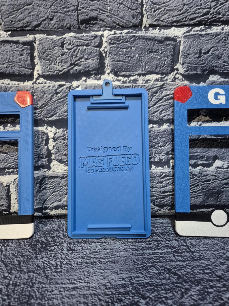 May include: A blue 3D-printed clipboard with a clip at the top and the text "Designed By MAS FUEGO 3D PRODUCTIONS" printed on the front. The clipboard is flanked by two blue frames with red and white accents.