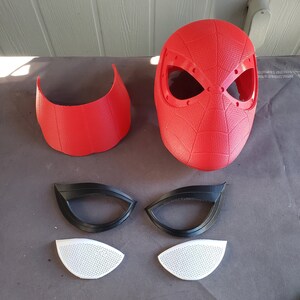 May include: A red and black Spider-Man mask with a textured surface. The mask is made up of five pieces: a main mask, two eye pieces, and two smaller pieces for the eyes. The eye pieces are black and the smaller pieces are white.