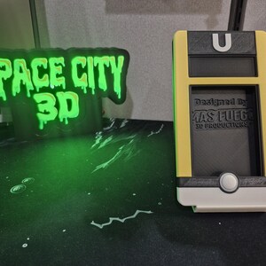 May include: A yellow, black, and white 3D-printed phone holder with the text "Designed by MAS FUEGO 3D PRODUCTIONS." A neon green sign with the text "SPACE CITY 3D" is in the background.