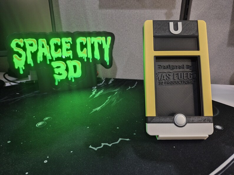 May include: A yellow, black, and white 3D-printed phone holder with the text "Designed by MAS FUEGO 3D PRODUCTIONS." A neon green sign with the text "SPACE CITY 3D" is in the background.