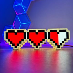 May include: Pixelated heart-shaped light fixture with three hearts, two red and one half-white. The light has a black outline and a beige frame. The background is a gradient of blue and purple.