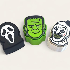 May include: Three Halloween-themed decorative items. A black, oval-shaped item with a white ghost face, a green Frankenstein's monster head, and a white and black clown face. The items are likely made of a hard material.