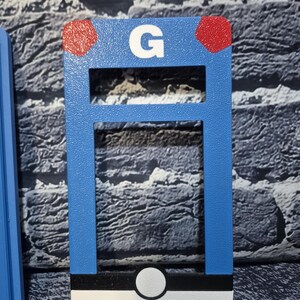 May include: A blue rectangular object with a white "G" and two red dots. The object has a white, black, and white design at the bottom. The object is a decorative item.