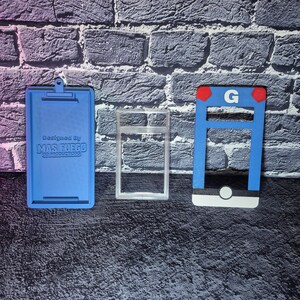 May include: Three 3D-printed objects: a blue clipboard with text, a clear rectangular frame, and a blue and white structure with a red accent and the letter "G". The objects are on a textured surface.