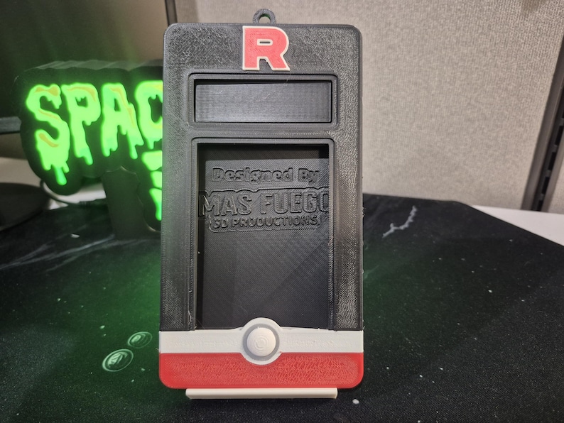 May include: Black and red rectangular case with a white and red accent at the bottom. The top has a red "R" and a rectangular opening. The text "Designed By MAS FUEGO 3D PRODUCTIONS" is visible. A green neon sign that says "SPACE" is in the background.