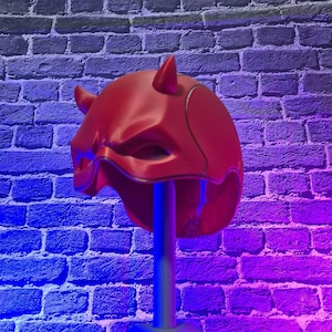 May include: A red superhero mask with two small horns, displayed on a silver stand. The mask has a sleek design with eye cutouts and a defined mouth area. The background is a brick wall illuminated with blue and purple lighting.