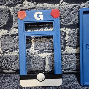 May include: A blue, rectangular 3D-printed item with a white "G" and red accents. The item has a black and white stripe at the bottom with a white circular button. The item is a holder for a game.