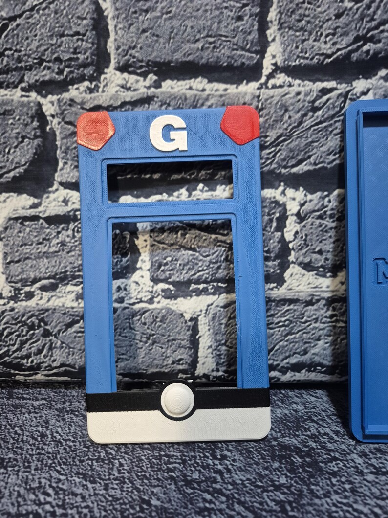 May include: A blue, rectangular 3D-printed item with a white "G" and red accents. The item has a black and white stripe at the bottom with a white circular button. The item is a holder for a game.