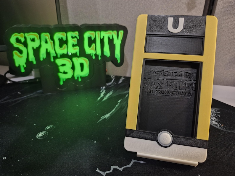 May include: A yellow and black 3D-printed phone case with a white base and a circular button. The case has the letter "U" on top and the text "Designed By MAS FUEGO 3D PRODUCTIONS" on the front. A glowing green "SPACE CITY 3D" sign is in the background.
