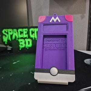 May include: A purple 3D-printed phone case featuring a white "M" on top, pink accents, and a black and white design at the bottom. The text "Designed by MAS FUEGO 3D PRODUCTIONS" is visible. The case is designed to hold a phone.