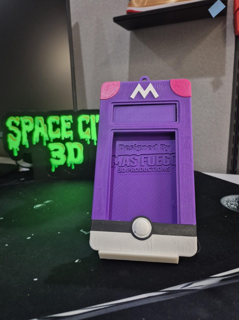 May include: A purple 3D-printed phone case featuring a white "M" on top, pink accents, and a black and white design at the bottom. The text "Designed by MAS FUEGO 3D PRODUCTIONS" is visible. The case is designed to hold a phone.