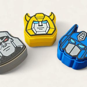 May include: Three colorful, character-themed boxes. One is yellow with a Bumblebee design, another is blue with an Optimus Prime design, and the third is gray with a Megatron design. The boxes are likely for storage or display.