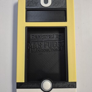 May include: A 3D-printed device in yellow, black, and white, featuring a U-shaped loop at the top. The design includes a rectangular window and a circular button. The text "Designed By MAS FUEGO 3D PRODUCTIONS" is visible.