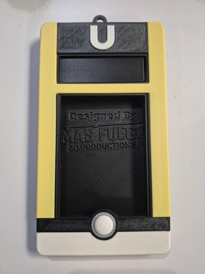 May include: A 3D-printed device in yellow, black, and white, featuring a U-shaped loop at the top. The design includes a rectangular window and a circular button. The text "Designed By MAS FUEGO 3D PRODUCTIONS" is visible.
