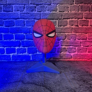 May include: A red and black Spider-Man mask with white eye lenses, displayed on a gray stand. The mask features a web pattern and is set against a brick wall illuminated with blue and red lighting.