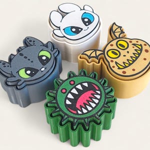 May include: Four colorful cookie cutters featuring dragon designs. The cutters include a gray dragon with green eyes, a white dragon with blue eyes, a yellow dragon with a toothy grin, and a green monster with a red mouth.