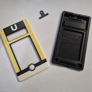 May include: A disassembled phone case with a yellow, black, and white design. The case has a rectangular shape with a circular button and the letter "U". The black base has the text "Designed by MAS FUEGO SD PRODUCTIONS".