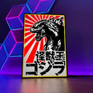 May include: A rectangular black and white Godzilla art print with a red sunburst background. The print features a detailed illustration of Godzilla and Japanese text. The print is framed with a yellow border.