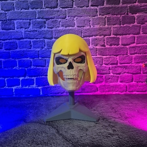 May include: A three-dimensional skull mask featuring a bright yellow bob-style wig with bangs. The skull is off-white with dark eye sockets and a detailed jawline. The mask is mounted on a gray triangular stand, positioned for display purposes.