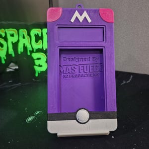 May include: A purple 3D-printed phone case with a white 'M' logo and pink accents. The case features a rectangular opening and a circular design at the bottom. Text on the case reads 'Designed By MAS FUEGO 3D PRODUCTIONS'.