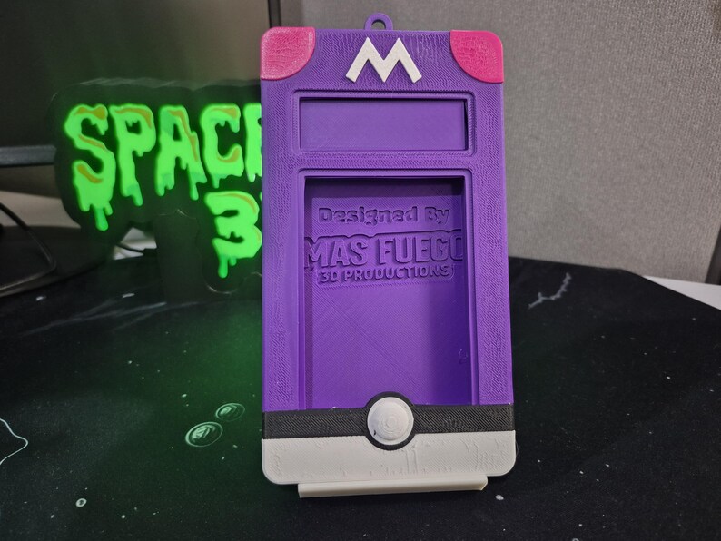 May include: A purple 3D-printed phone case with a white 'M' logo and pink accents. The case features a rectangular opening and a circular design at the bottom. Text on the case reads 'Designed By MAS FUEGO 3D PRODUCTIONS'.