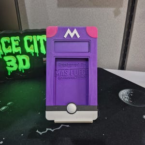 May include: A purple 3D-printed phone case with a white "M" on the top and pink accents. The case has a slot for a phone and a white button. The text "Designed By MAS FUEGO 3D PRODUCTIONS" is printed on the case.