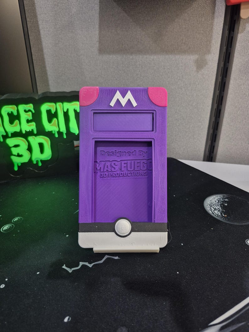 May include: A purple 3D-printed phone case with a white "M" on the top and pink accents. The case has a slot for a phone and a white button. The text "Designed By MAS FUEGO 3D PRODUCTIONS" is printed on the case.