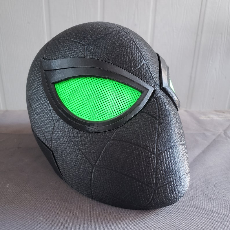 May include: A black and green 3D printed mask with a spider web design. The mask has two green eyes with a mesh pattern.