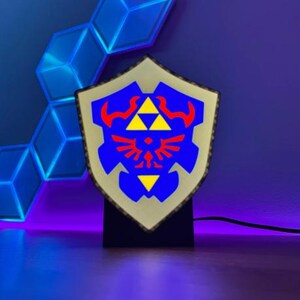 May include: A shield-shaped lamp with a blue, red, and yellow design, reminiscent of a video game shield. The lamp is mounted on a black base and emits a soft glow. The background features geometric blue light panels.