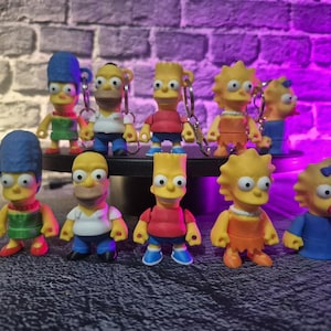 May include: A collection of ten Simpsons character figurines. The figures include Homer, Marge, Bart, Lisa, and Maggie Simpson. Each figure is brightly colored and has a keychain attached. The figures are standing on a black surface.