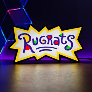 May include: A neon sign featuring the word "Rugrats" in a playful, stylized font. The sign has a white background with a yellow jagged border. The letters are purple with red, green, and yellow accents.