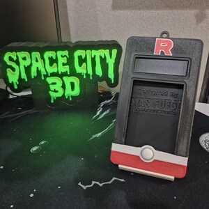 May include: A black and green 3D-printed sign that reads "SPACE CITY 3D" with a dripping green font. Next to it is a black and red device with the letter "R" on top and the text "Designed By MAS FUEGO 3D Productions".