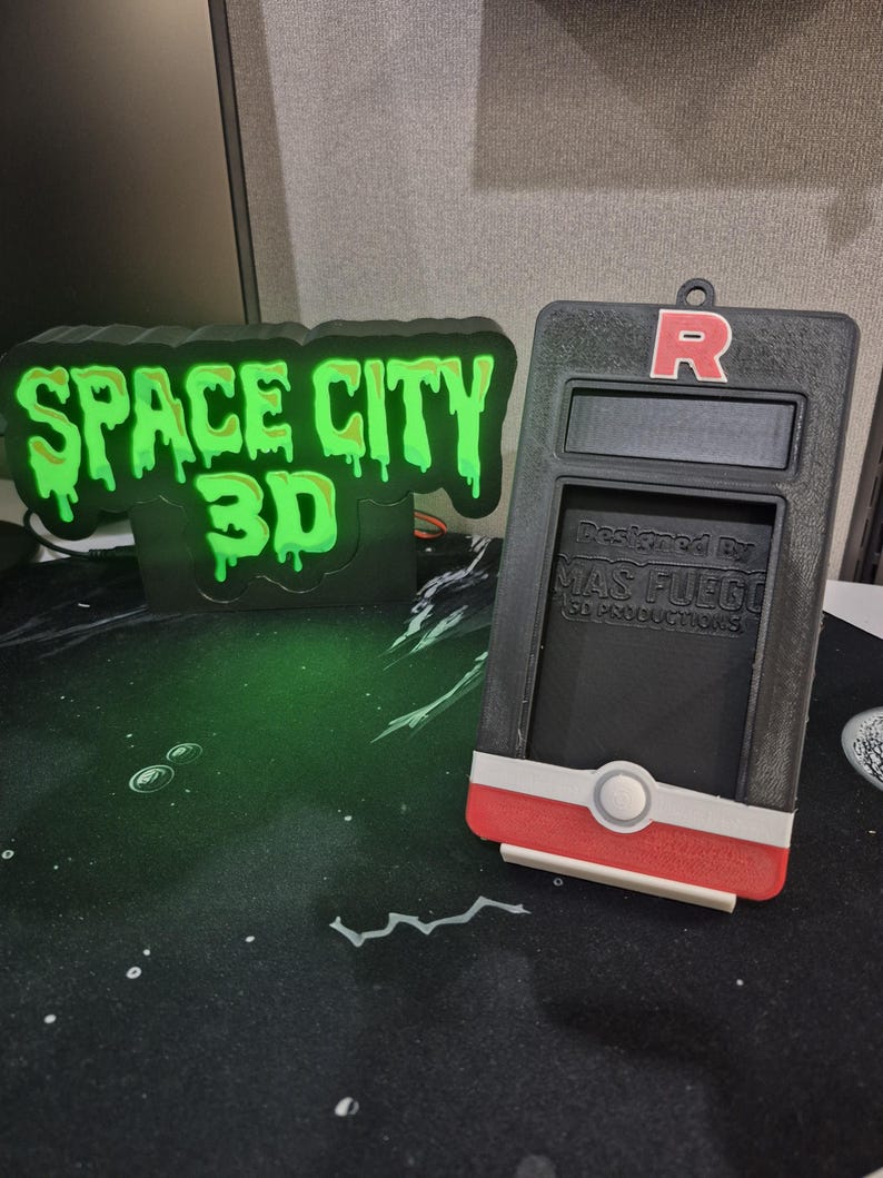 May include: A black and green 3D-printed sign that reads "SPACE CITY 3D" with a dripping green font. Next to it is a black and red device with the letter "R" on top and the text "Designed By MAS FUEGO 3D Productions".