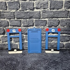 May include: Three blue 3D-printed items. Two have a rectangular frame with a white base, a black stripe, and a red accent. The third is a blue clipboard with the text "Designed By MAS FUEGO".