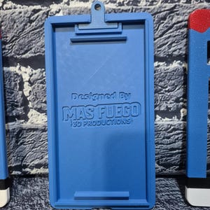 May include: A blue 3D-printed clipboard with a clip at the top and a bar at the bottom. The text "Designed By MAS FUEGO 3D PRODUCTIONS" is printed on the clipboard. The clipboard is on a textured surface.