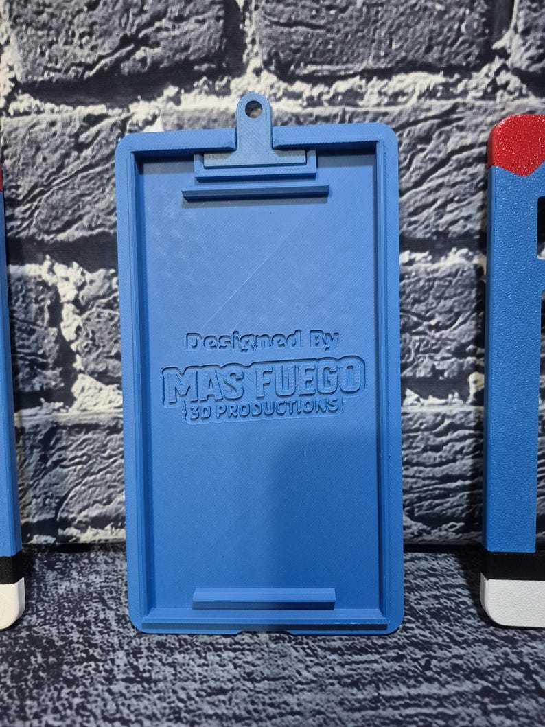 May include: A blue 3D-printed clipboard with a clip at the top and a bar at the bottom. The text "Designed By MAS FUEGO 3D PRODUCTIONS" is printed on the clipboard. The clipboard is on a textured surface.
