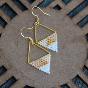 May include: A pair of gold triangle earrings with white, peach, and gold beads. The earrings are hanging from a wooden surface.